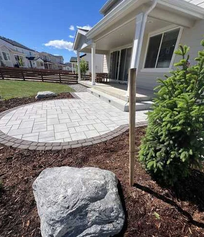 Curved pale paver patio beside a covered porch, landscape boulder and evergreen tree.