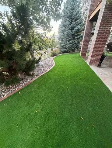 Artificial turf curving around a shaded side yard between a home and planting beds.