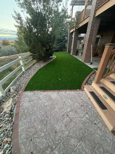Turf beneath a raised deck connected to a textured concrete patio and rock borders.