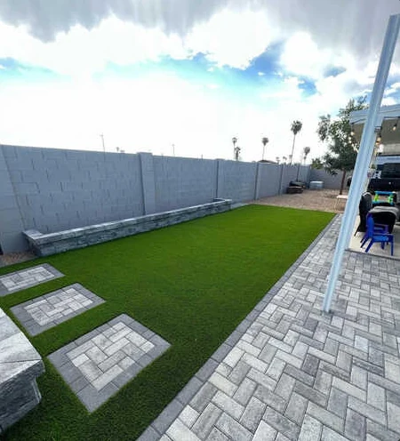 Artificial turf lawn with square paver stepping stones, gray patio and a raised stone border.